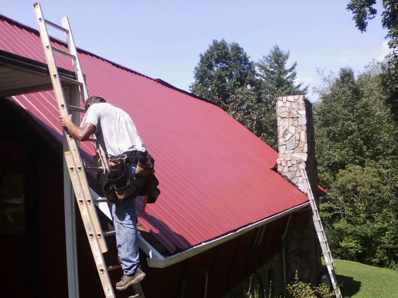 Roofer on a ladder beside a red metal roof and stone chimney — Soffit & Fascia Repair by Roofing Brothers in Beavertown